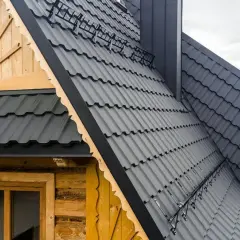 Dach mansardowy konstrukcja z ciemnoszarej blachy. Drewniana ściana z oknem i ozdobnymi elementami. W tle zielone wzgórza.