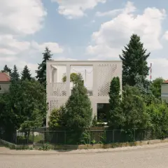 Nowoczesny dom typu kostka z białej cegły, otoczony zielenią. Widać fragmenty sąsiednich budynków i błękitne niebo z chmurami.