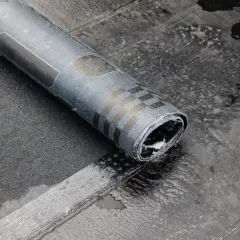 Rolka papy na zimno leży na mokrej powierzchni dachu. Widać fragmenty czarnego materiału i wzory na papie.