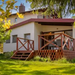 Bungalow z drewnianym tarasem i schodkami, otoczony zielonym ogrodem i kwitnącym drzewem.