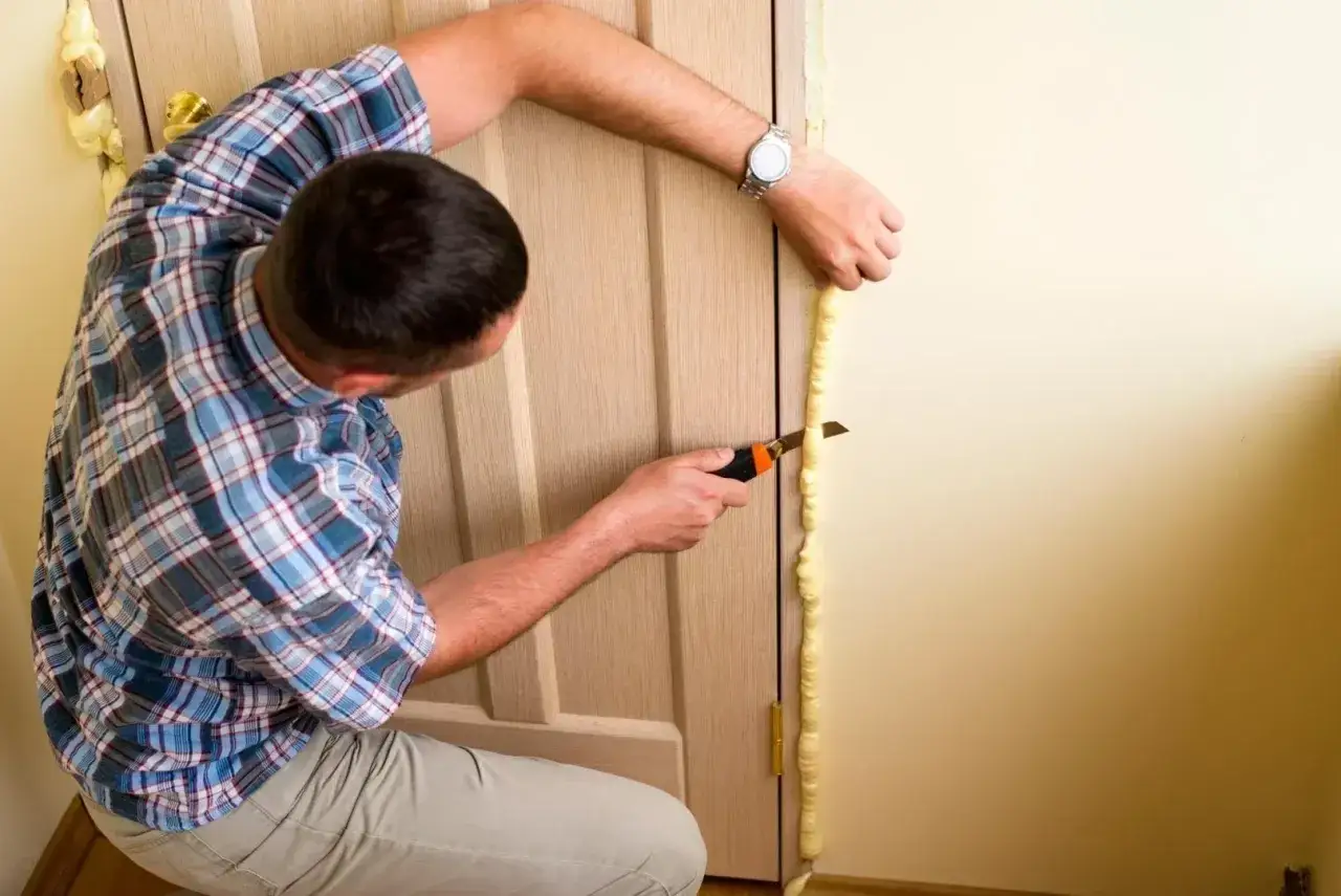 Mężczyzna przycina piankę montażową wok&oacute;ł drzwi. Zastanawiasz się, ile za wstawienie drzwi?