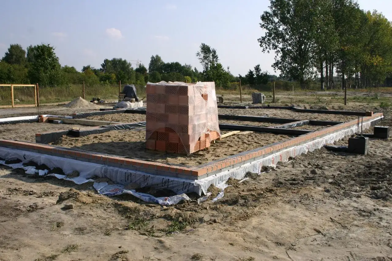 Fundamenty budynku w stanie surowym, z paletą cegieł przykrytą folią. Widać zarys ścian fundamentowych i przygotowany teren.