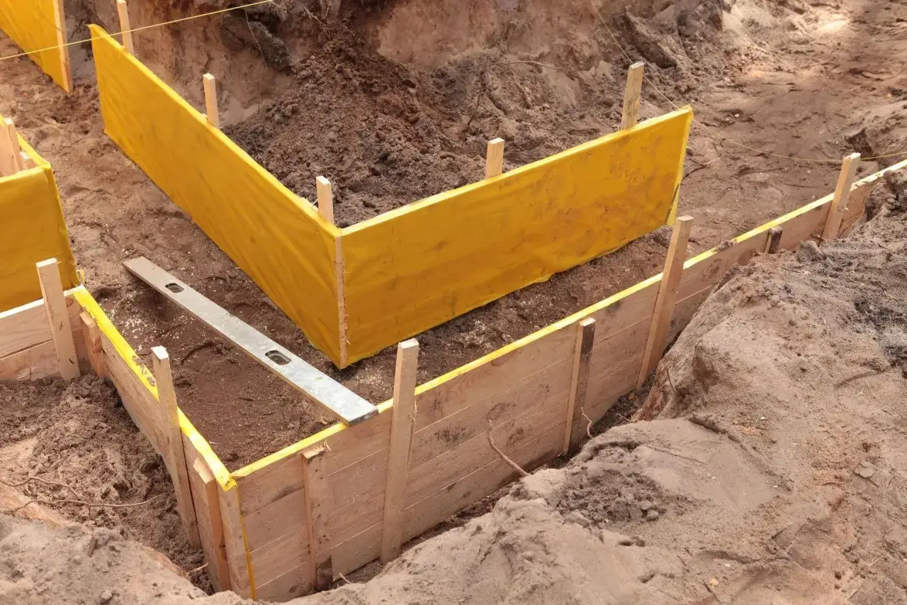 Wykonano wykopy pod fundamenty. Widać drewniane szalunki i żółtą folię. Poziomica leży w wykopie.